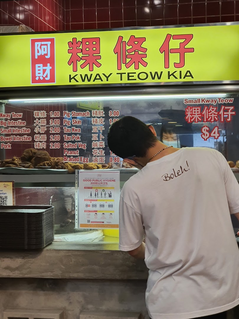 A food stall counter displaying a menu for Kway Teow Kia, with a customer at the counter.
