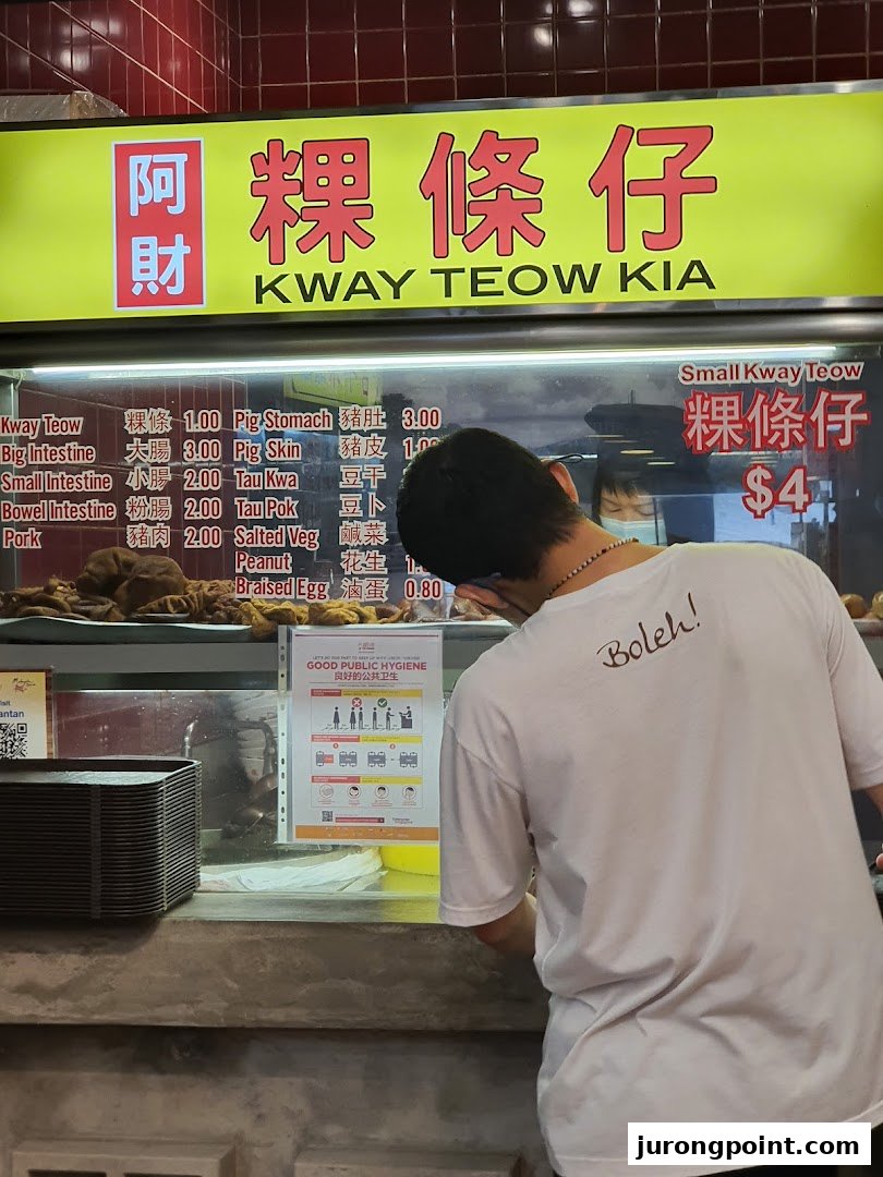 A food stall counter displaying a menu for Kway Teow Kia, with a customer at the counter.