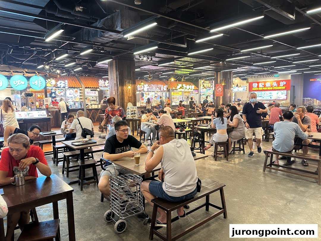 A lively food court filled with patrons enjoying diverse Malaysian cuisine at various stalls.