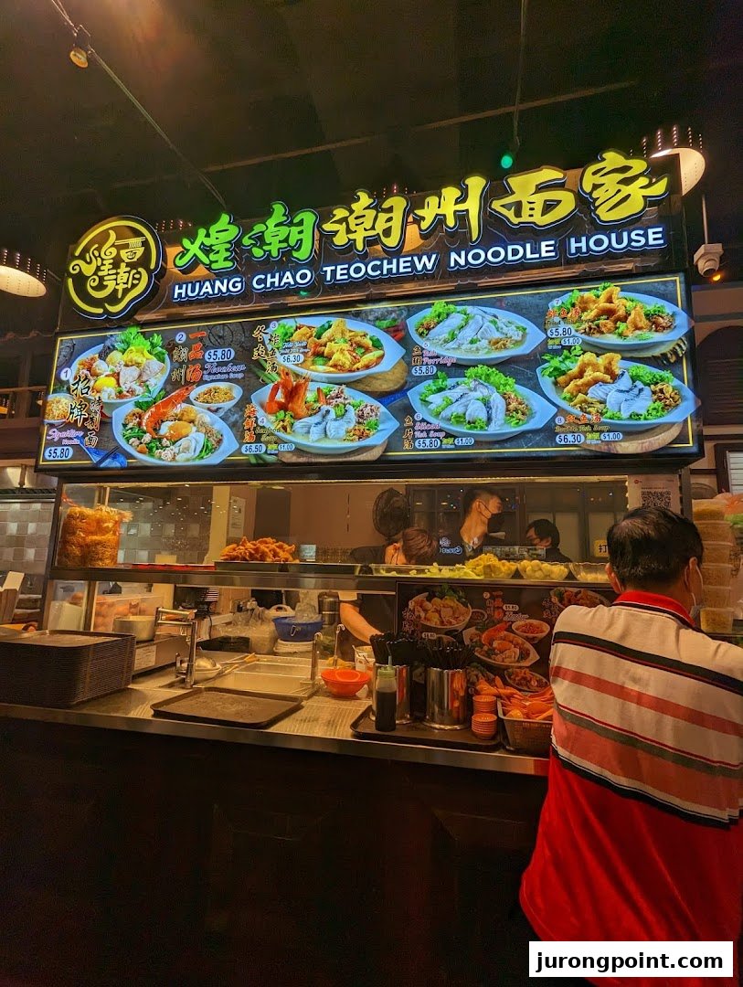 A vibrant food stall counter with a large illuminated menu board showcasing various Teochew noodle dishes.