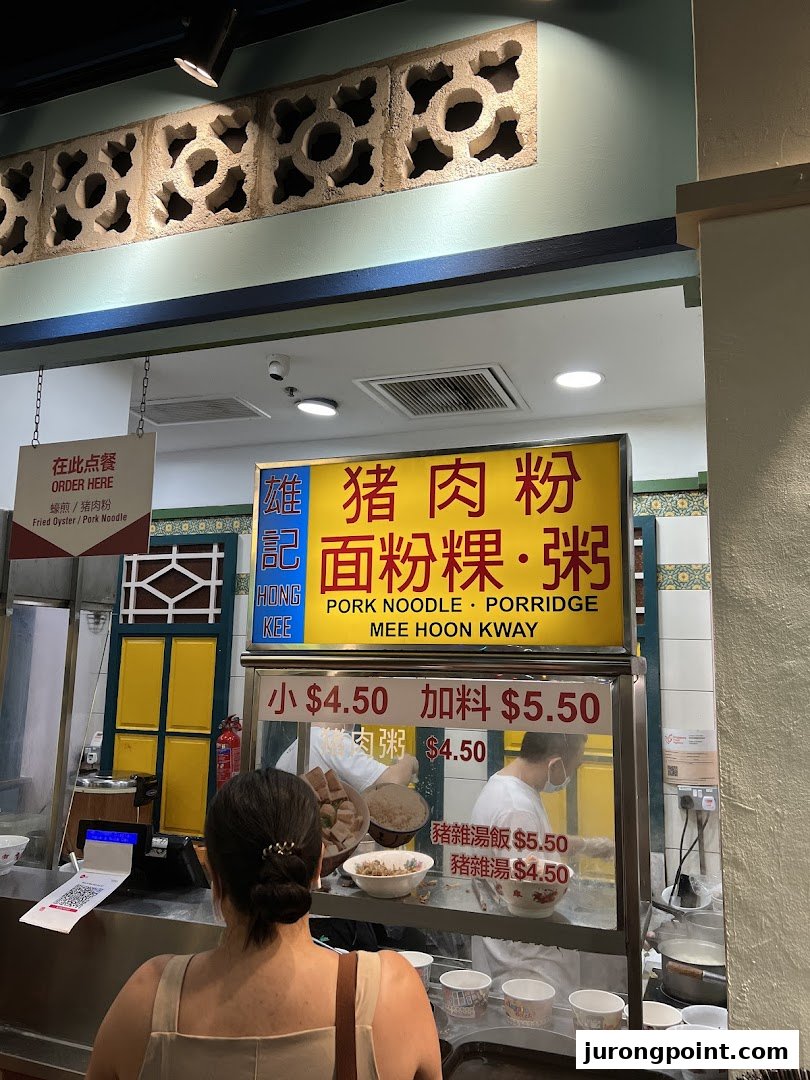 A customer at a food stall counter with a menu board displaying pork noodle and porridge options.