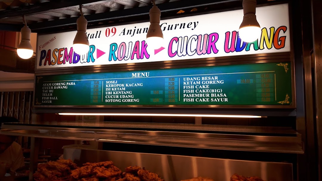 A brightly lit menu board displaying various Malaysian fritters, snacks, and dishes with prices.