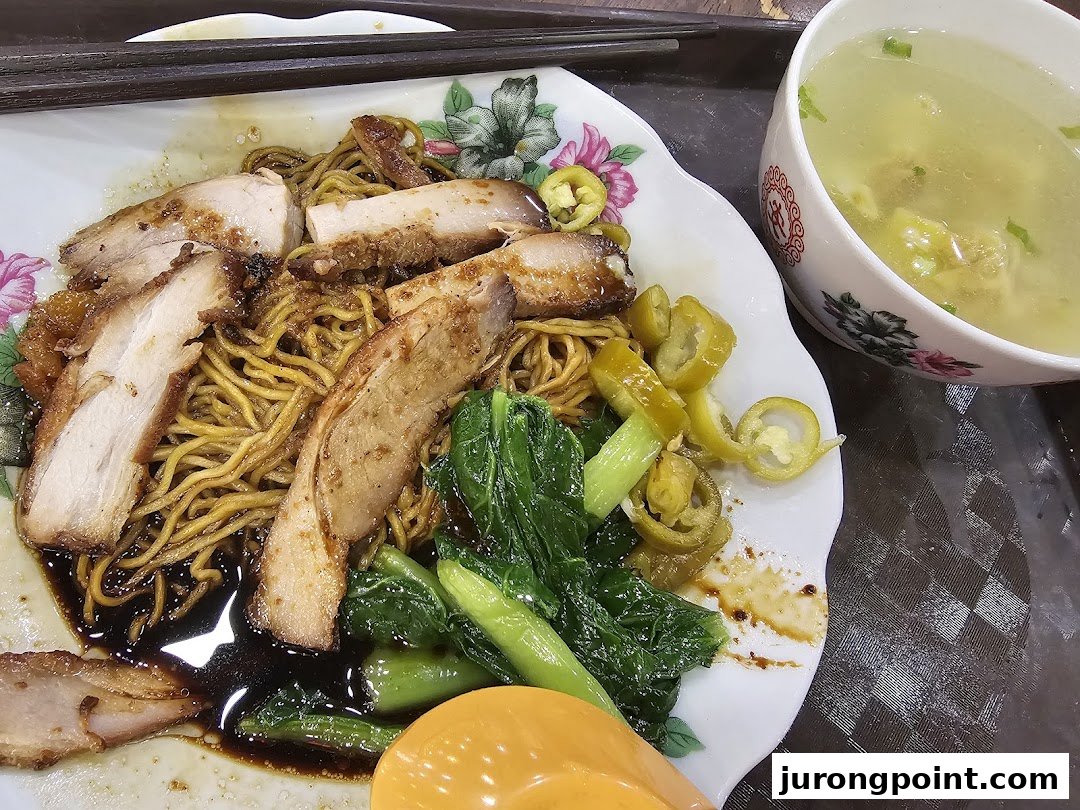 A delicious plate of wanton mee with char siew, green vegetables, pickled chilies, and a side of soup.