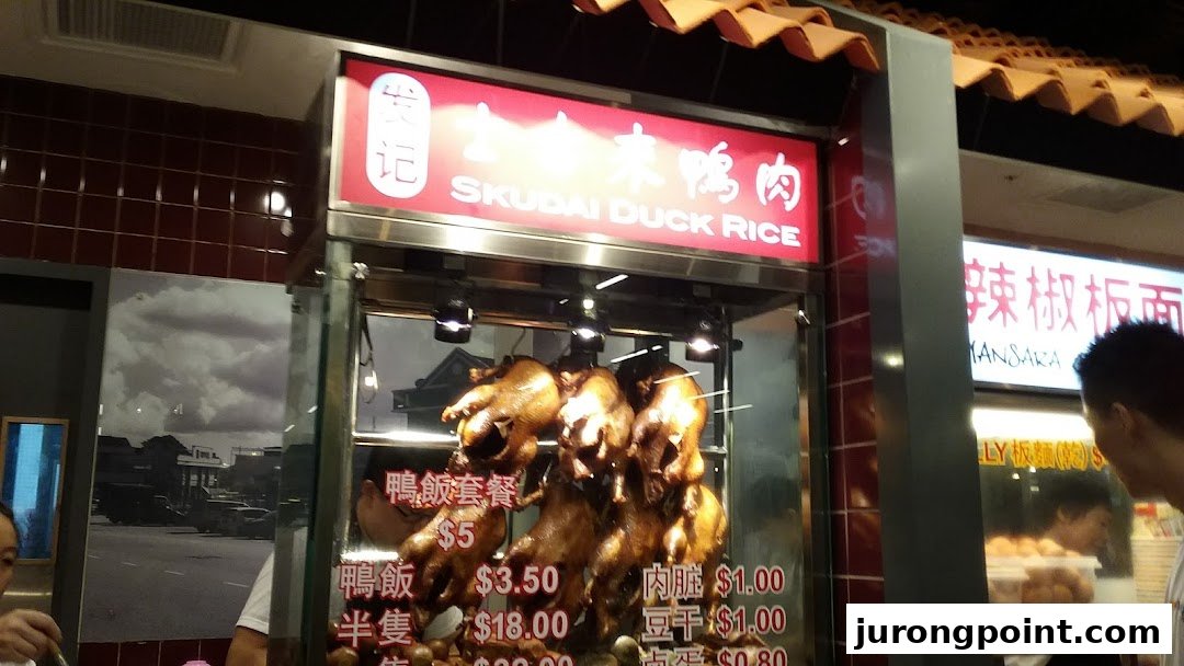 A food stall displaying roasted ducks and a menu with prices for duck rice and side dishes.