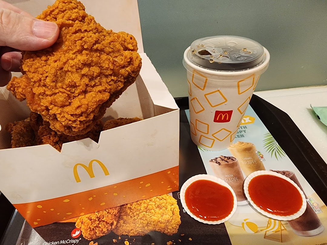 A hand holds a piece of crispy fried chicken from a McDonald's box, alongside a drink and sauces.