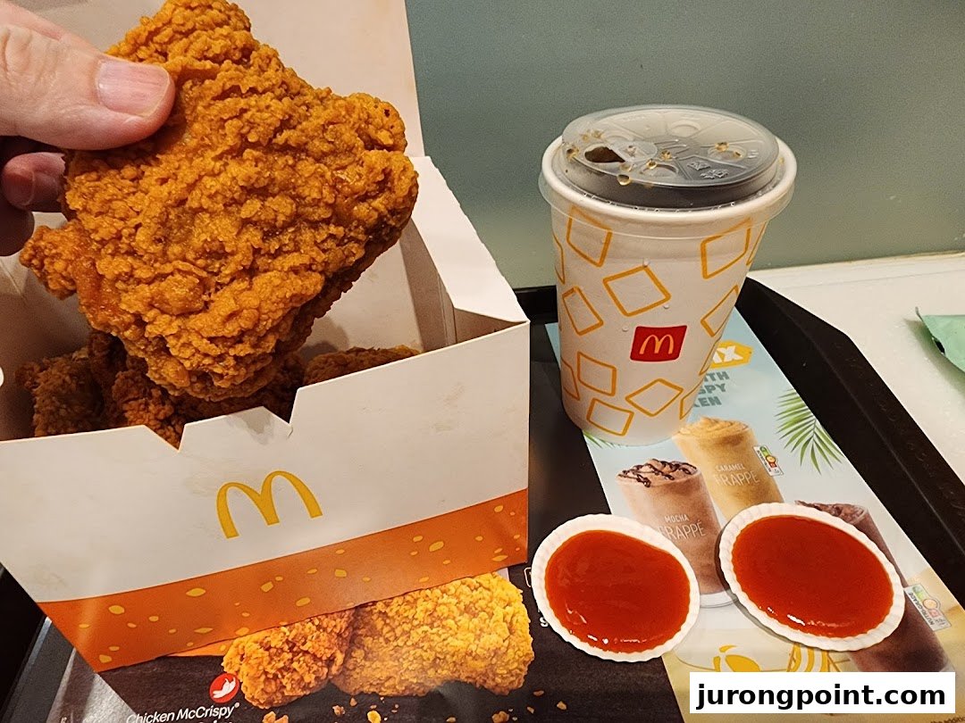 A hand holds a piece of crispy fried chicken from a McDonald's box, alongside a drink and sauces.