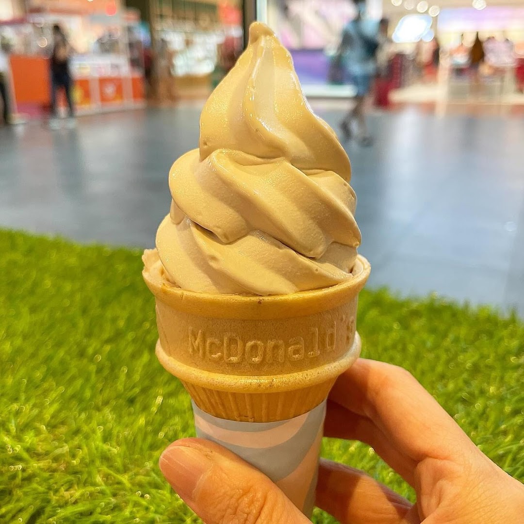 A hand holds a McDonald's soft-serve ice cream cone with the logo visible, set against a mall background.