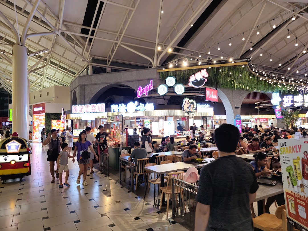 A bustling food court with various food stalls, diners, and a lively atmosphere under a high ceiling.