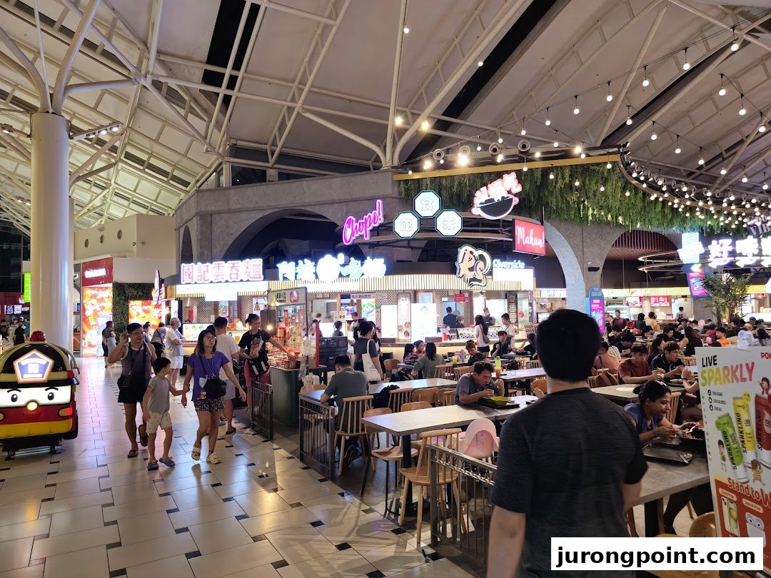 A bustling food court with various food stalls, diners, and a lively atmosphere under a high ceiling.