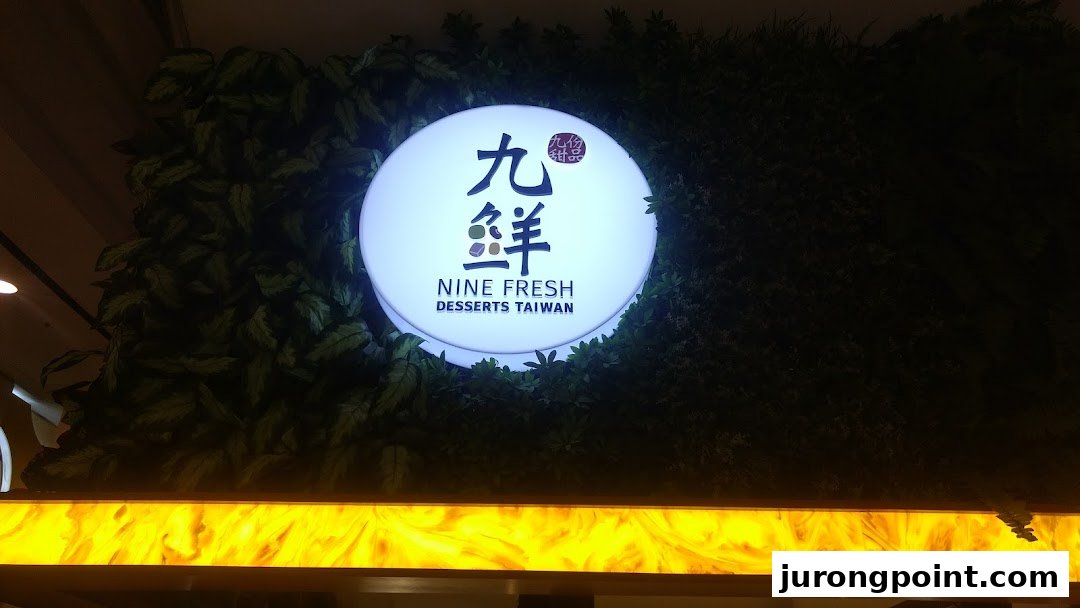 An illuminated circular sign for Nine Fresh Desserts Taiwan set against a green artificial foliage wall.