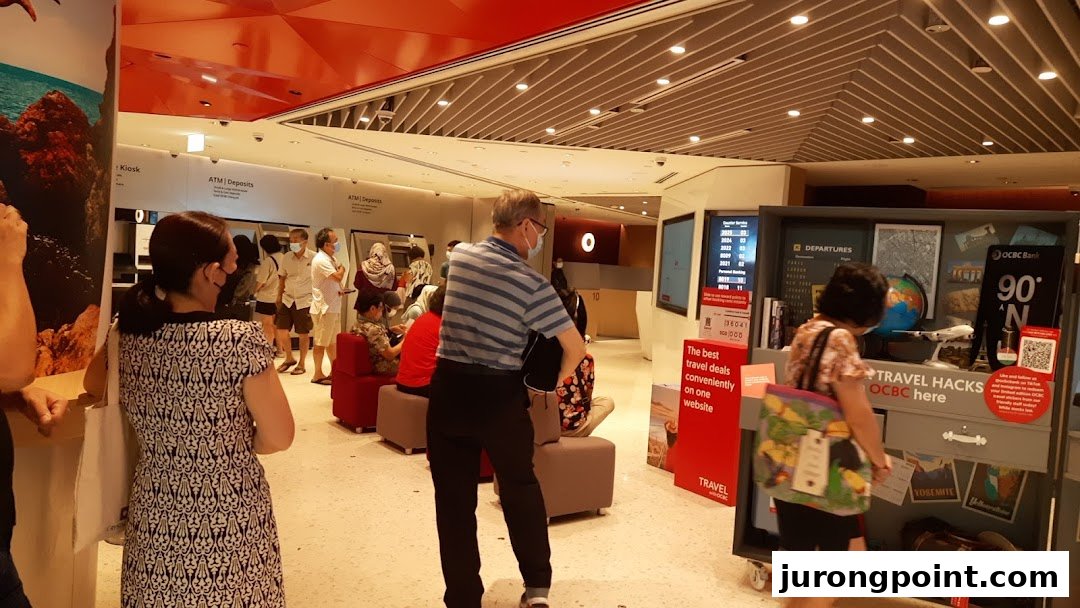 Customers inside OCBC Bank Jurong Point, featuring ATMs and a travel-themed promotional display.