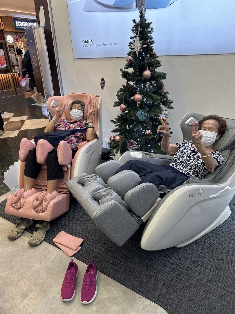 Two women enjoy OGAWA massage chairs, giving peace signs, next to a Christmas tree display.