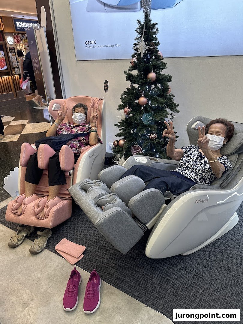 Two women enjoy OGAWA massage chairs, giving peace signs, next to a Christmas tree display.
