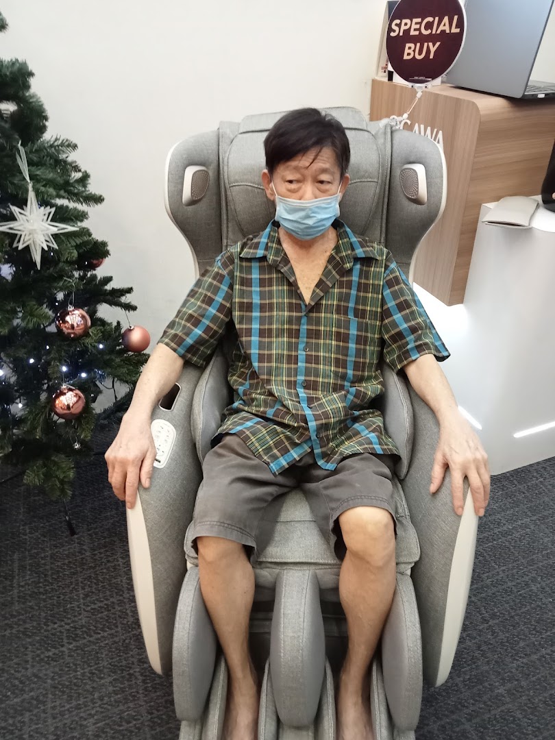 A man in a mask tests a grey massage chair at OGAWA, with a Christmas tree nearby.