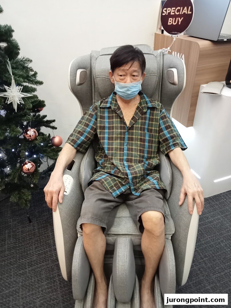 A man in a mask tests a grey massage chair at OGAWA, with a Christmas tree nearby.