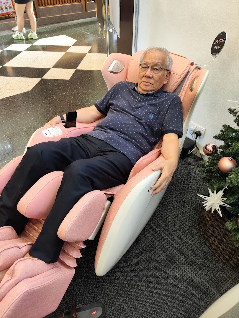 A man relaxing in a pink Ogawa massage chair, with a 'Special Deal' sign and Christmas tree nearby.