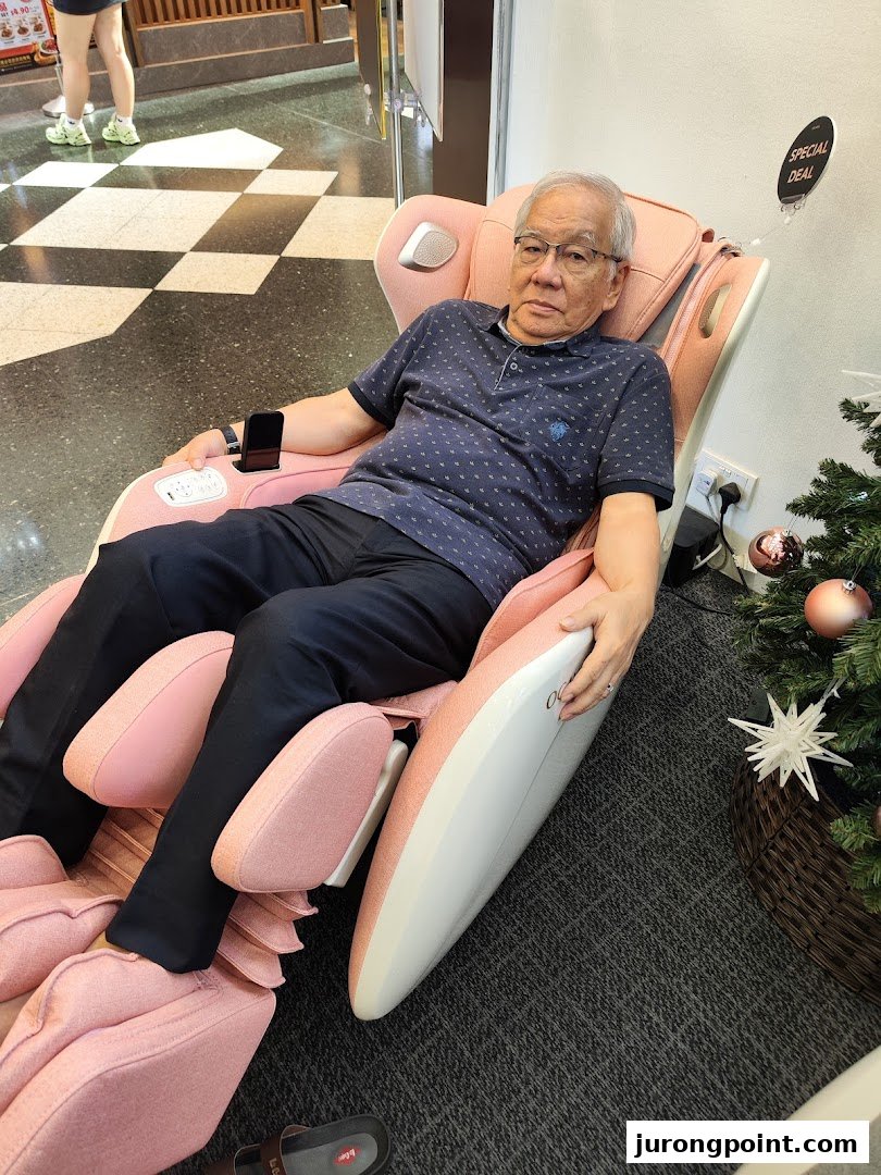 A man relaxing in a pink Ogawa massage chair, with a 'Special Deal' sign and Christmas tree nearby.