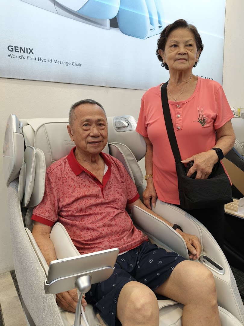 An elderly man relaxing in an OGAWA massage chair while an elderly woman stands beside him.