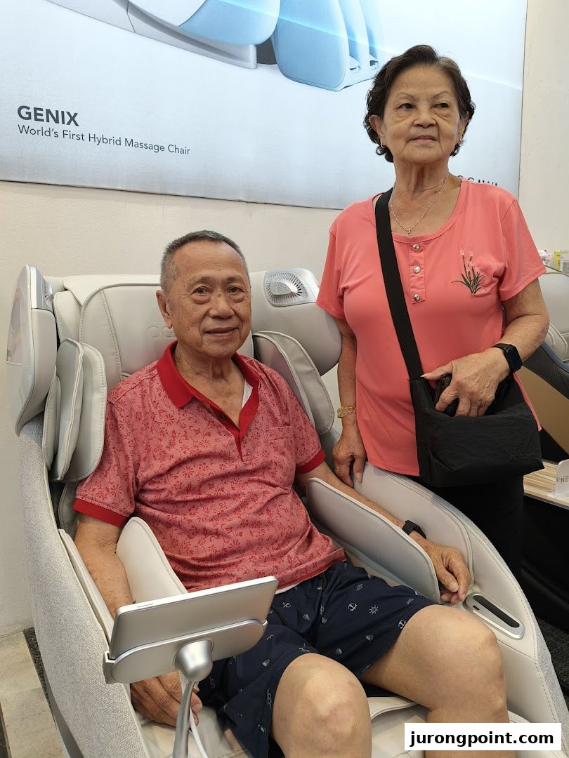 An elderly man relaxing in an OGAWA massage chair while an elderly woman stands beside him.