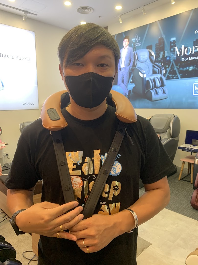 A person wearing a neck massager inside an OGAWA store, with massage chairs and promotional displays in the background.