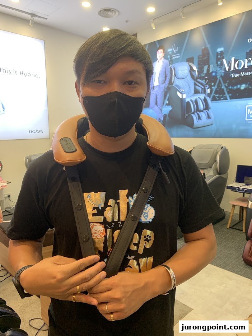 A person wearing a neck massager inside an OGAWA store, with massage chairs and promotional displays in the background.