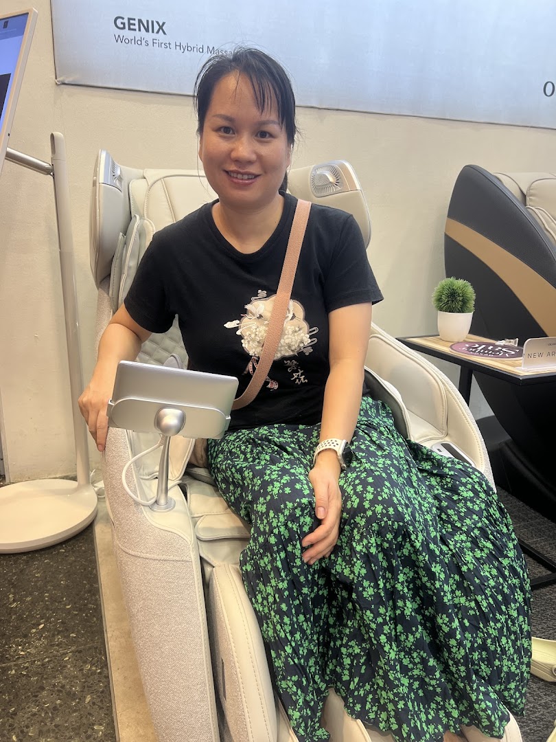 A woman smiling while trying out a massage chair at OGAWA, featuring a 'GENIX' model.