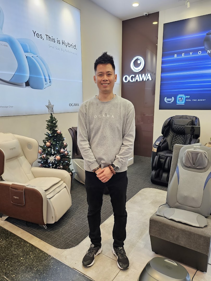 A smiling man stands amidst various massage chairs and a festive Christmas tree inside an OGAWA store.