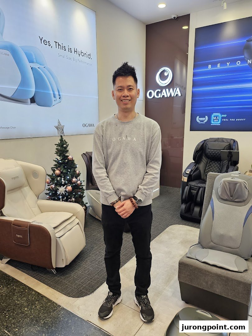 A smiling man stands amidst various massage chairs and a festive Christmas tree inside an OGAWA store.