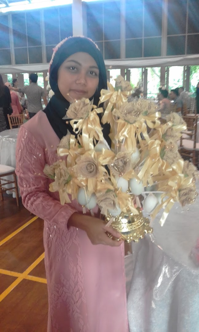 A woman in a pink dress and hijab holds a decorative golden and beige centerpiece at an indoor event.