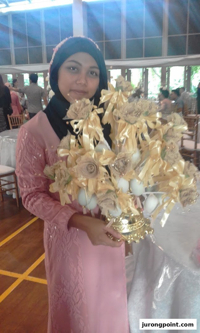 A woman in a pink dress and hijab holds a decorative golden and beige centerpiece at an indoor event.