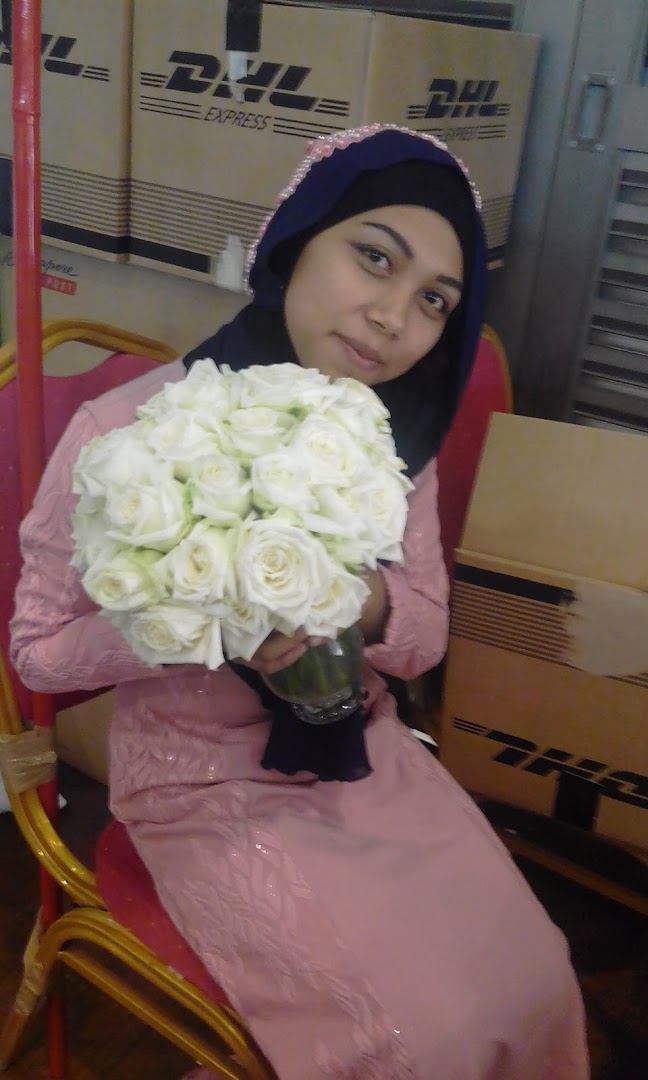 A woman in a pink dress and hijab holds a bouquet of white roses, with DHL boxes in the background.