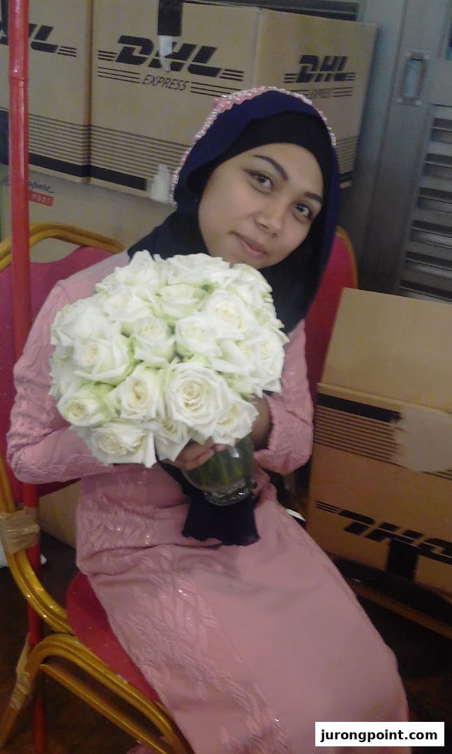 A woman in a pink dress and hijab holds a bouquet of white roses, with DHL boxes in the background.