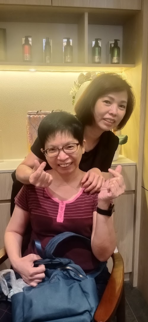 Two smiling women, one making a heart gesture, inside Oriental Hair Solutions with products on display.