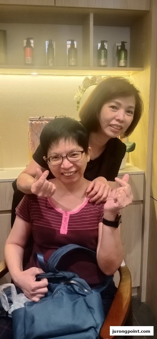 Two smiling women, one making a heart gesture, inside Oriental Hair Solutions with products on display.