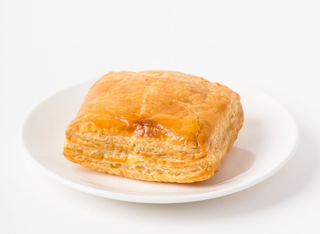 A golden-brown, flaky puff pastry sits on a clean white plate against a bright background.