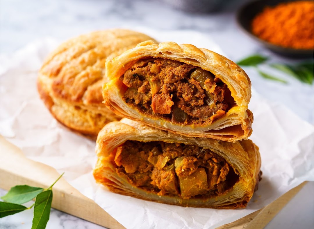 A close-up of a golden-brown pastry, likely a curry puff, cut open to reveal a savory, spiced filling.