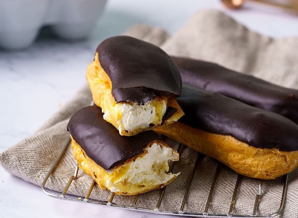 Several chocolate-covered eclairs filled with cream, resting on a metal cooling rack.