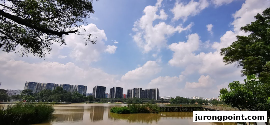 A serene lake with distant city buildings under a bright, cloudy sky, framed by lush trees.