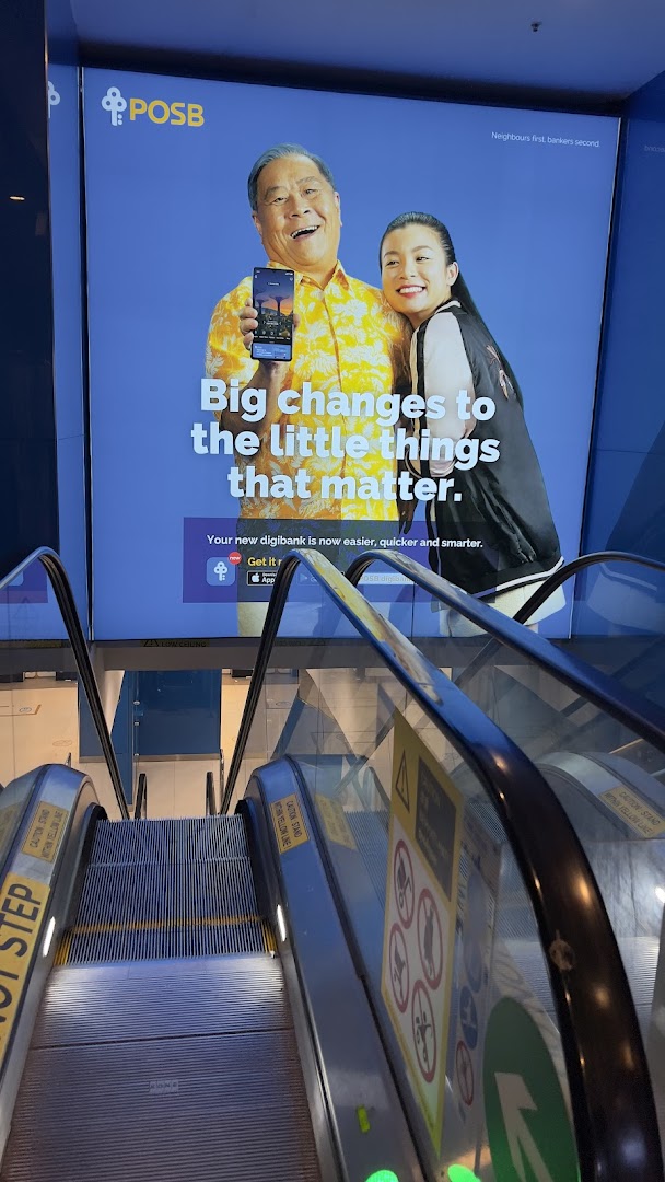 An escalator descends towards a large digital advertisement for POSB bank, featuring two smiling individuals.