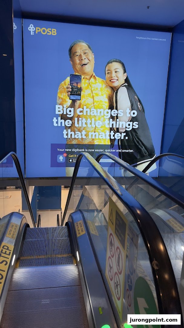 An escalator descends towards a large digital advertisement for POSB bank, featuring two smiling individuals.