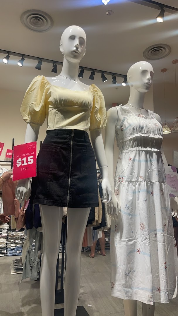 Two mannequins display trendy clothing, including a yellow puff-sleeve top, black skirt, and a white patterned dress, with a sale sign.