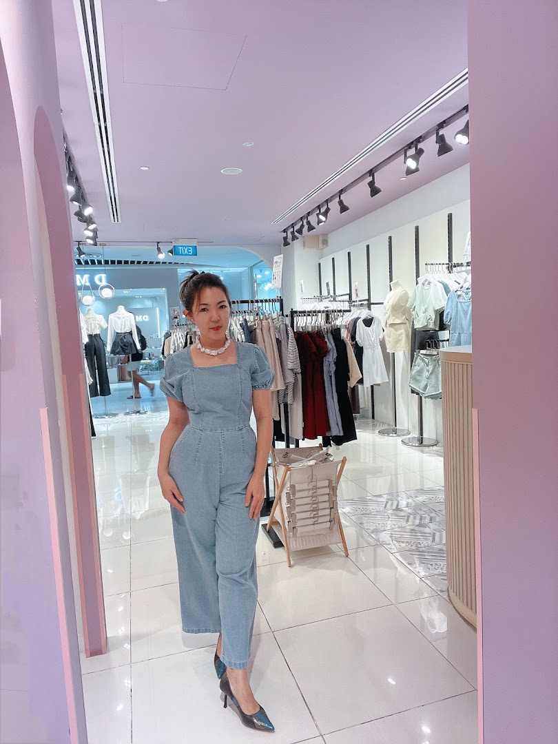 A woman in a denim jumpsuit poses inside Purpur, a brightly lit clothing boutique with racks of apparel.