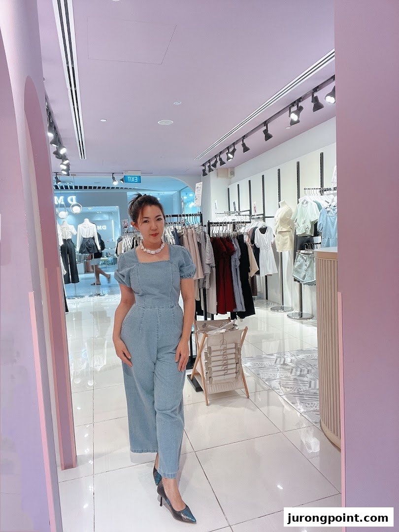 A woman in a denim jumpsuit poses inside Purpur, a brightly lit clothing boutique with racks of apparel.