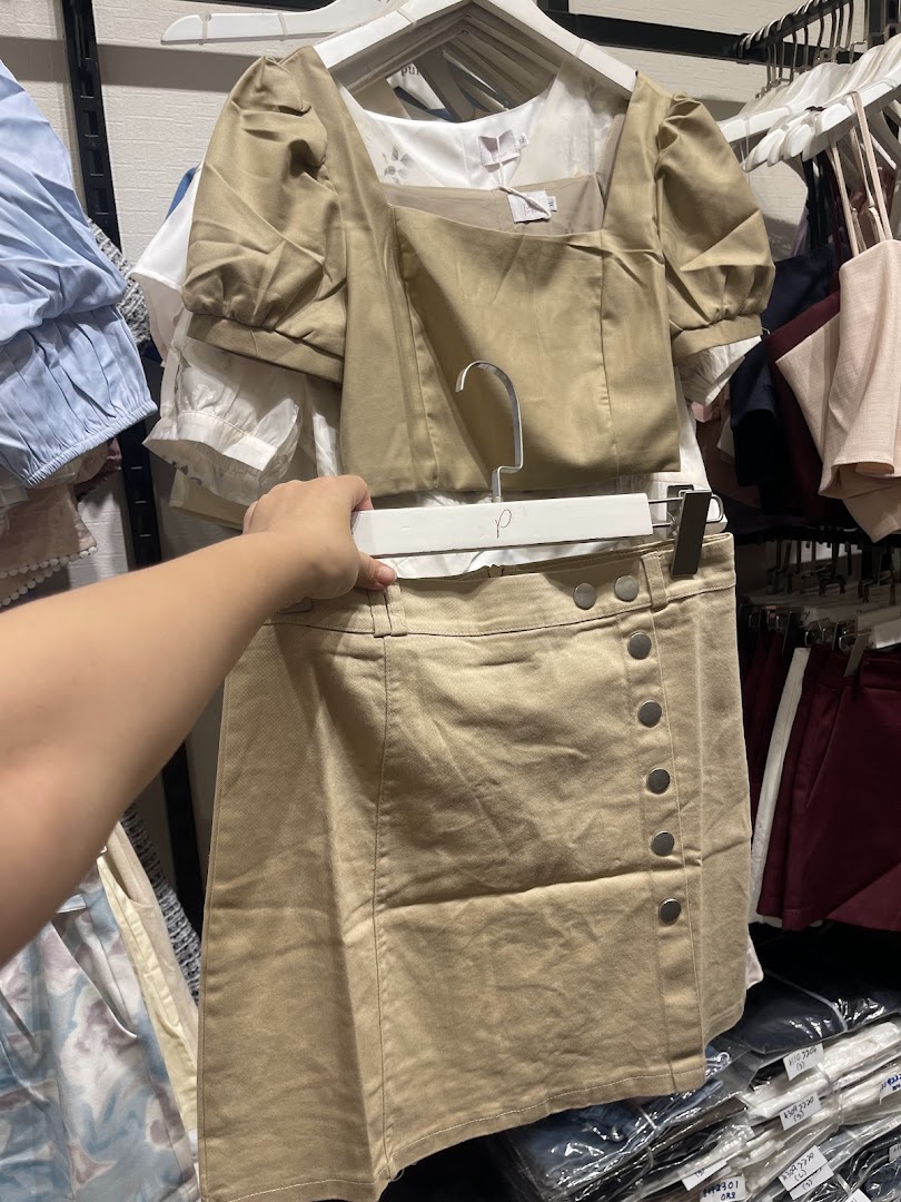 A hand holds up a stylish beige top and matching skirt set inside a clothing store.