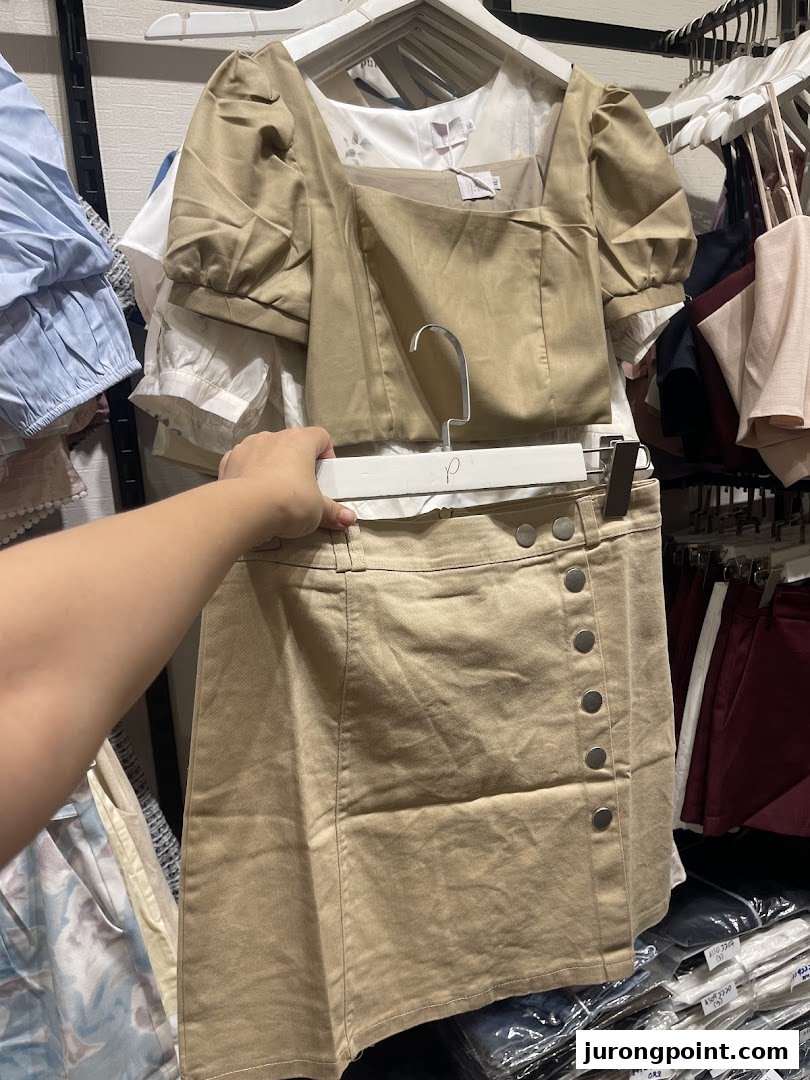 A hand holds up a stylish beige top and matching skirt set inside a clothing store.