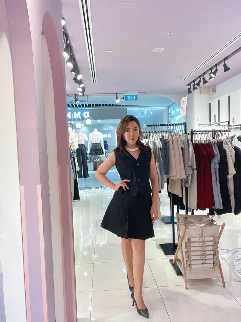 A woman poses inside a brightly lit clothing boutique with racks of fashionable apparel.