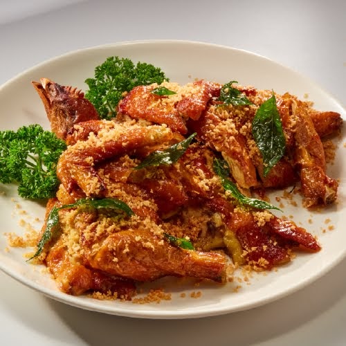 A golden-brown fried chicken dish, garnished with crispy crumbs and fresh herbs, served on a white plate.