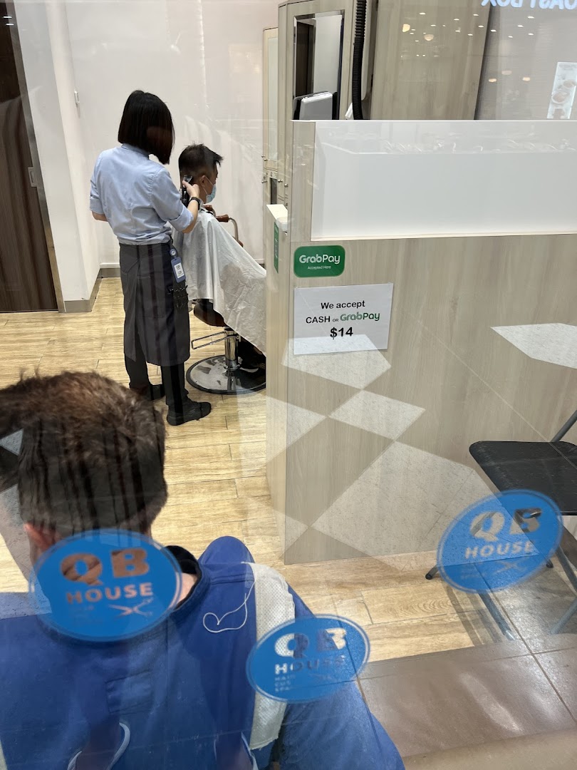 A stylist cuts a customer's hair inside QB House, viewed through a glass partition.