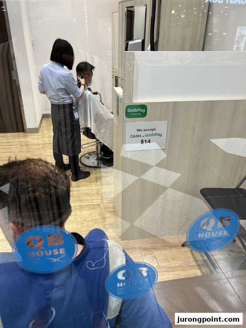 A stylist cuts a customer's hair inside QB House, viewed through a glass partition.