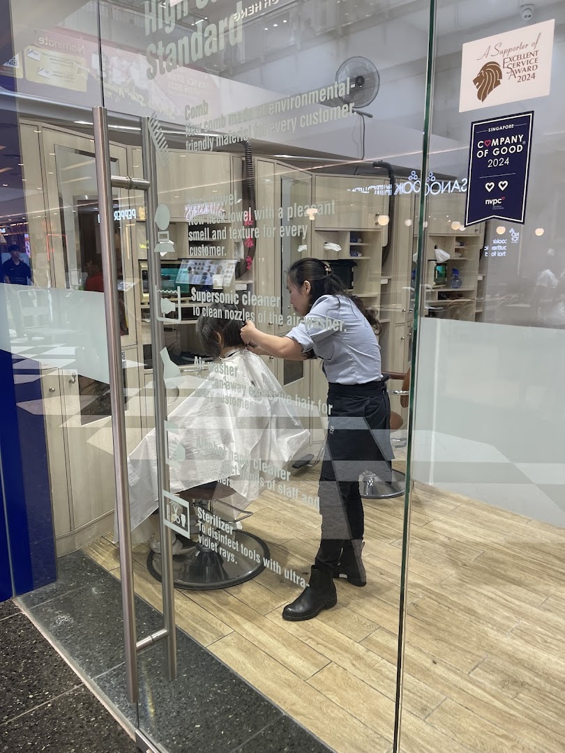 A stylist cuts a customer's hair inside QB House, visible through the glass entrance.